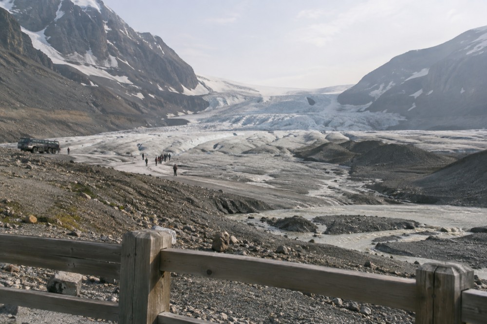 A gravel outwash plain leading toward the fenced edge of a glacier toe with bare rock beside the ice