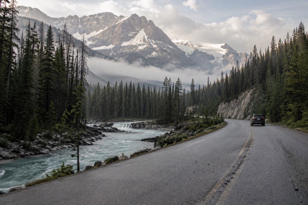A quiet asphalt road winding past a turquoise river between tall Rocky Mountain peaks