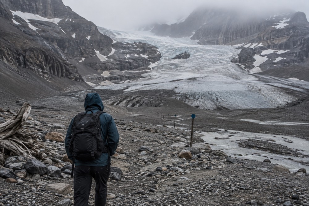 A hiker standing at the toe of the Athabasca Glacier beside recession marker signs