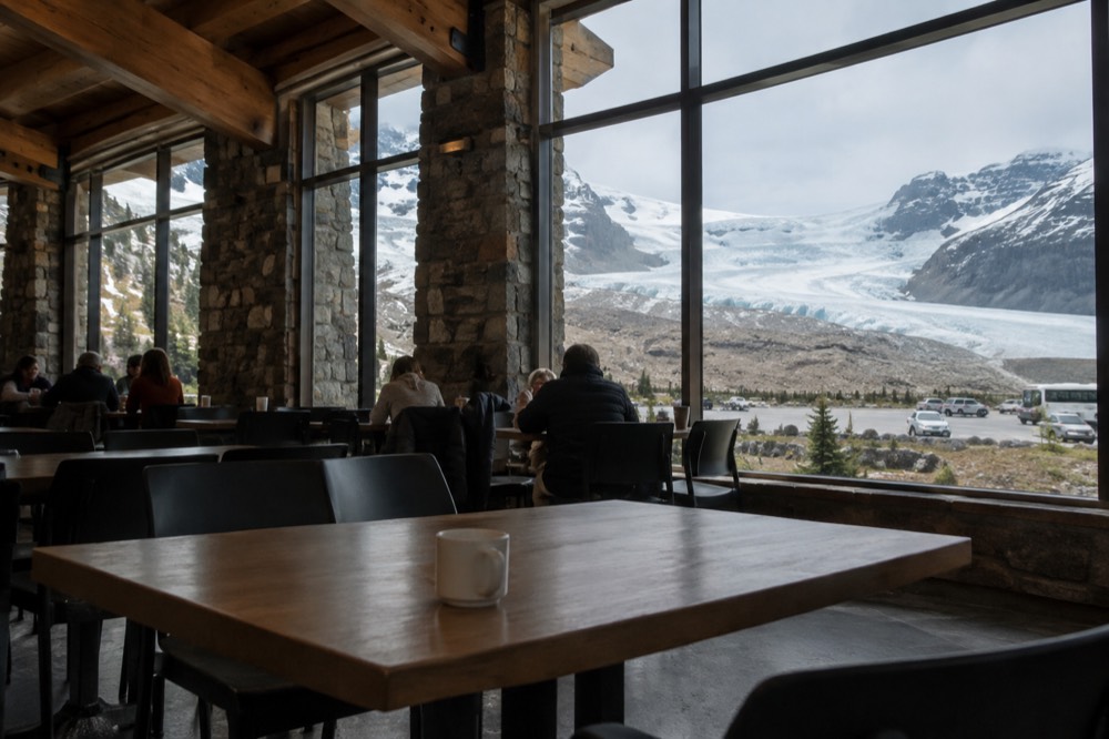 A dining area with large windows facing a snow-covered glacier across a mountain highway