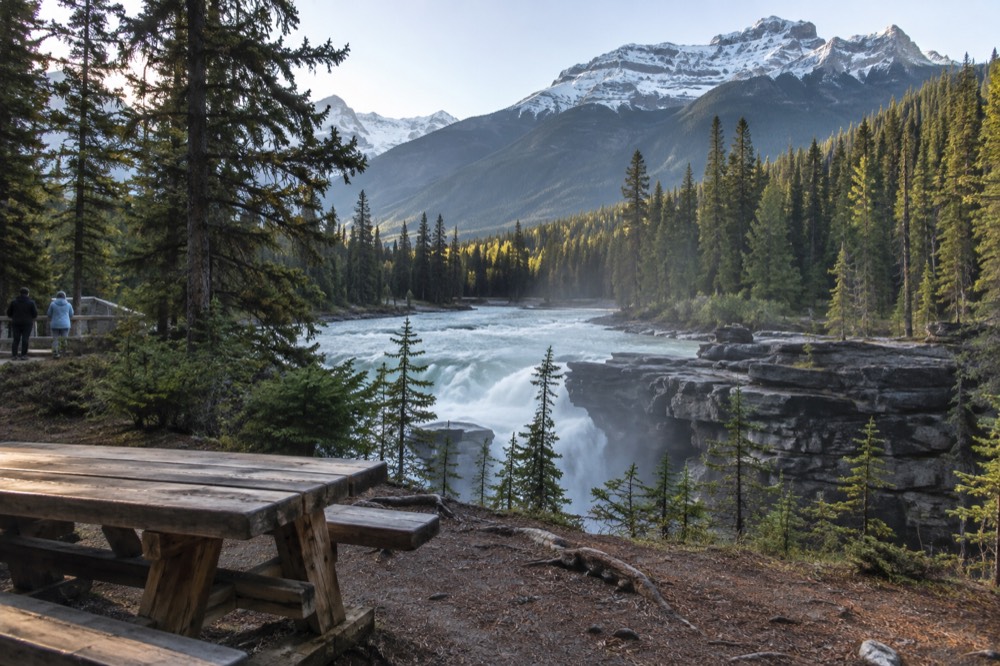 A roadside picnic table beside a turquoise river with tall snow-capped peaks behind it