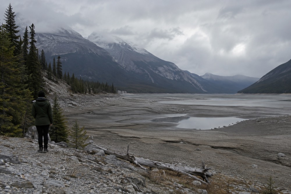 A long mountain lake ringed by forest with a gravel shoreline under clouds