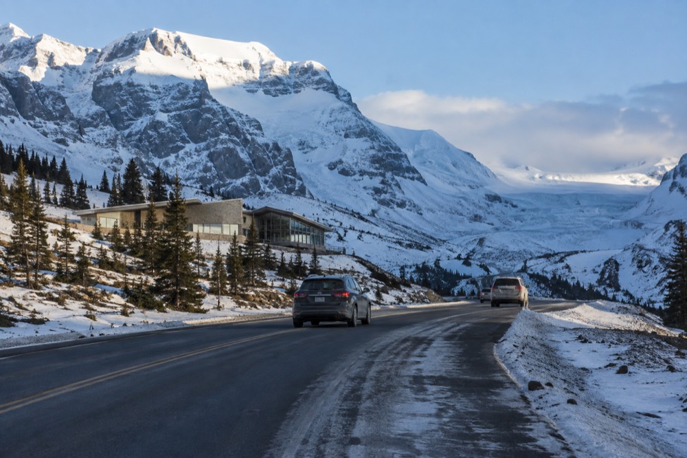 A two-lane mountain road curving between tall snow-covered peaks with a glacier in the distance