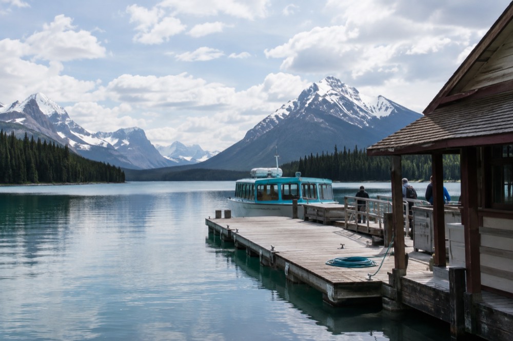 A wide glacial lake reflecting steep forested mountains under scattered clouds with a rocky shoreline