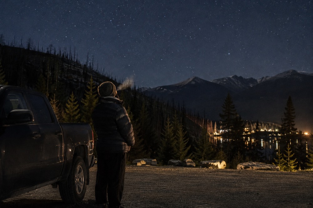 A person in a warm coat standing beside a parked car looking up at stars