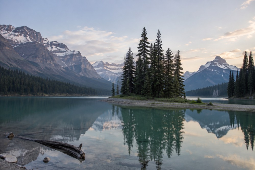 A small island with a few pine trees reflected perfectly in the still green water of a mountain lake, with snowy peaks in the background